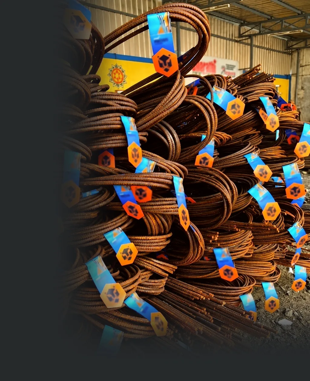Rust-brown coils of steel bars in a yard, each with a blue tag and an orange hexagon tag