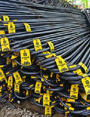 Yellow identification tags fixed along bundles of long TMT steel bars in a yard.