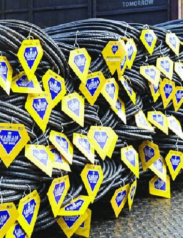 Yellow and blue identification tags hung on coiled TMT steel bars in a stockyard.